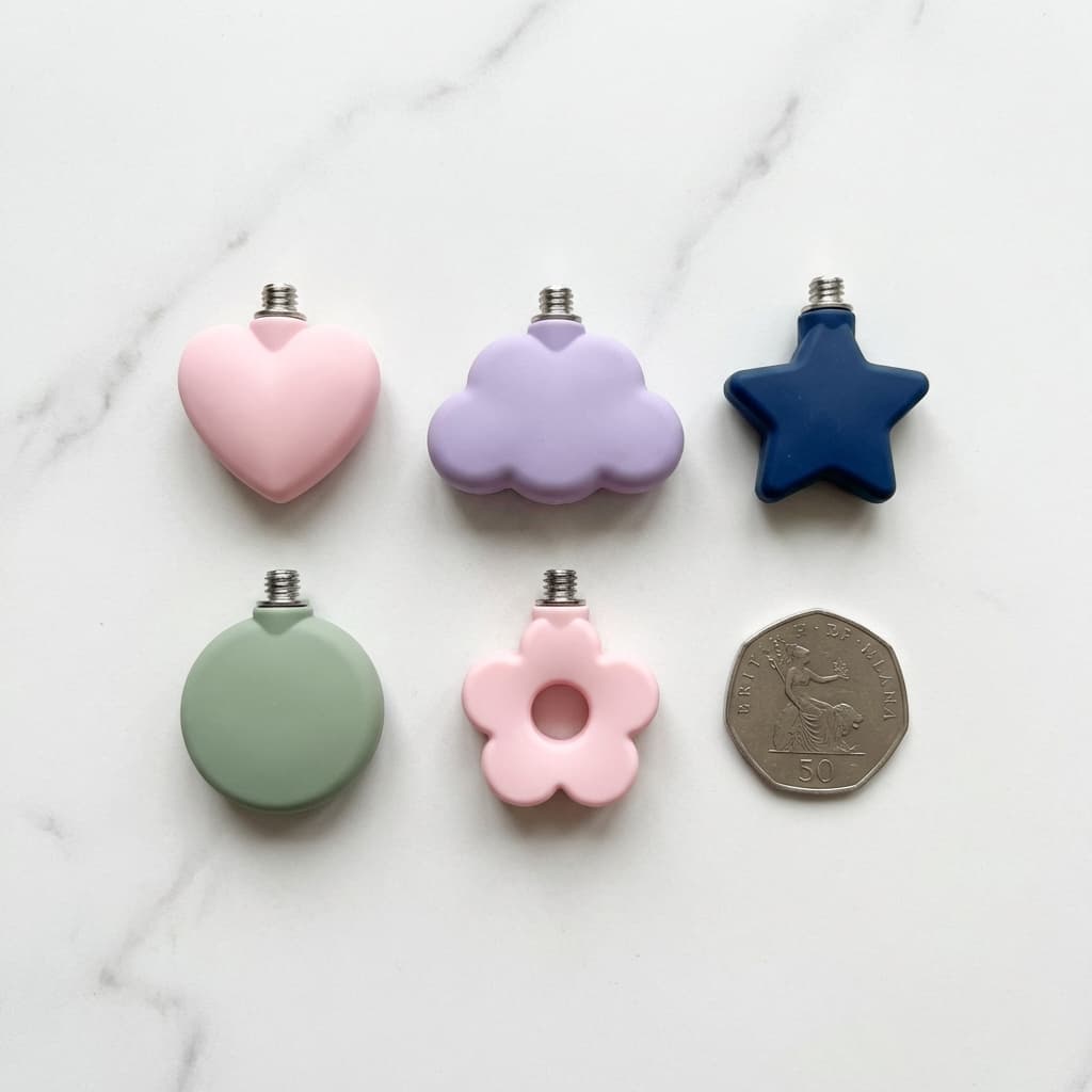 Five small silicone charms on a marble surface: a pink heart, a lilac cloud, a navy star, a green circle and a pink flower, next to a 50p coin for size